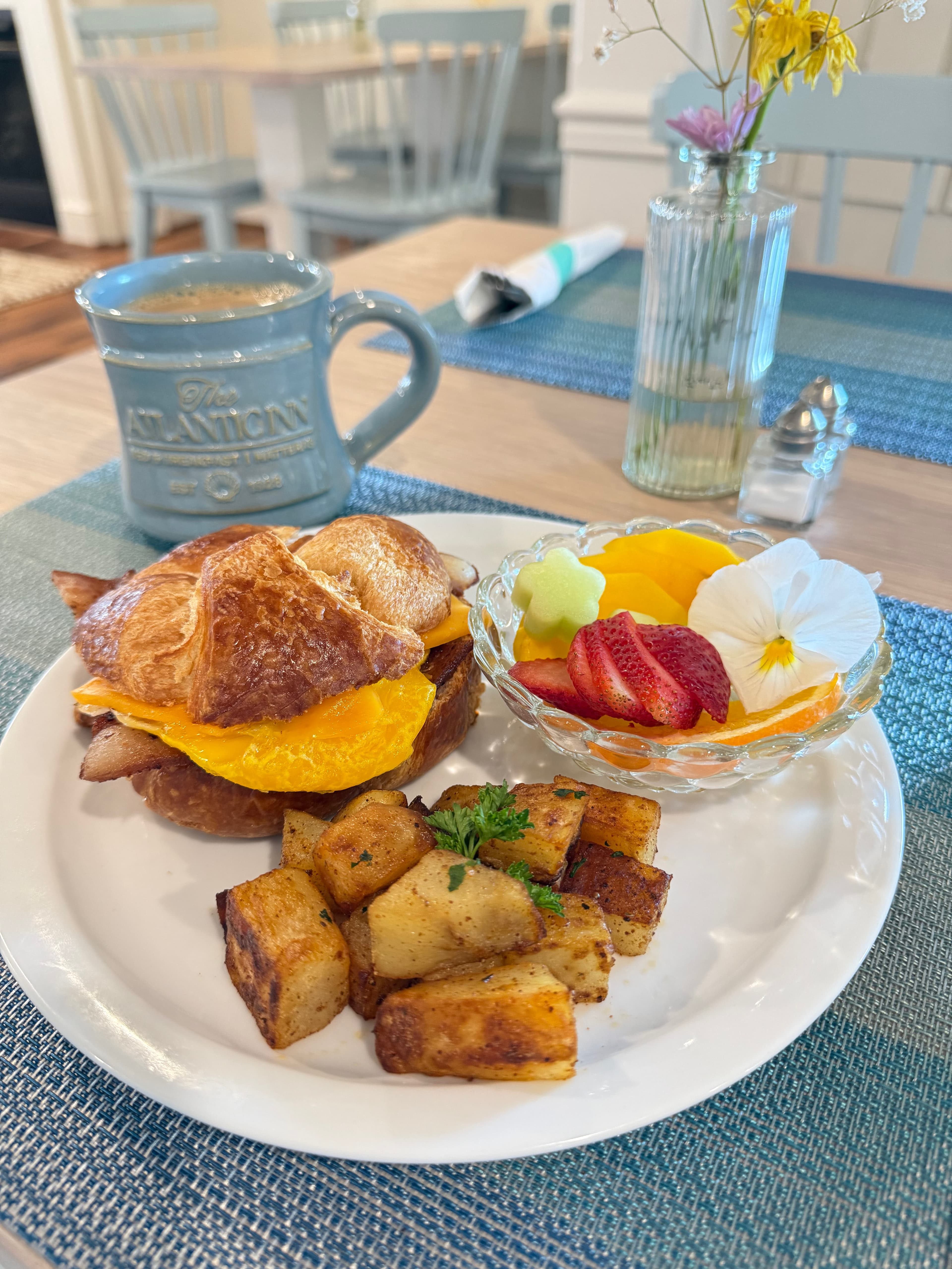 A plate with a croissant sandwich, roasted potatoes, and a fruit salad, accompanied by a mug of coffee.