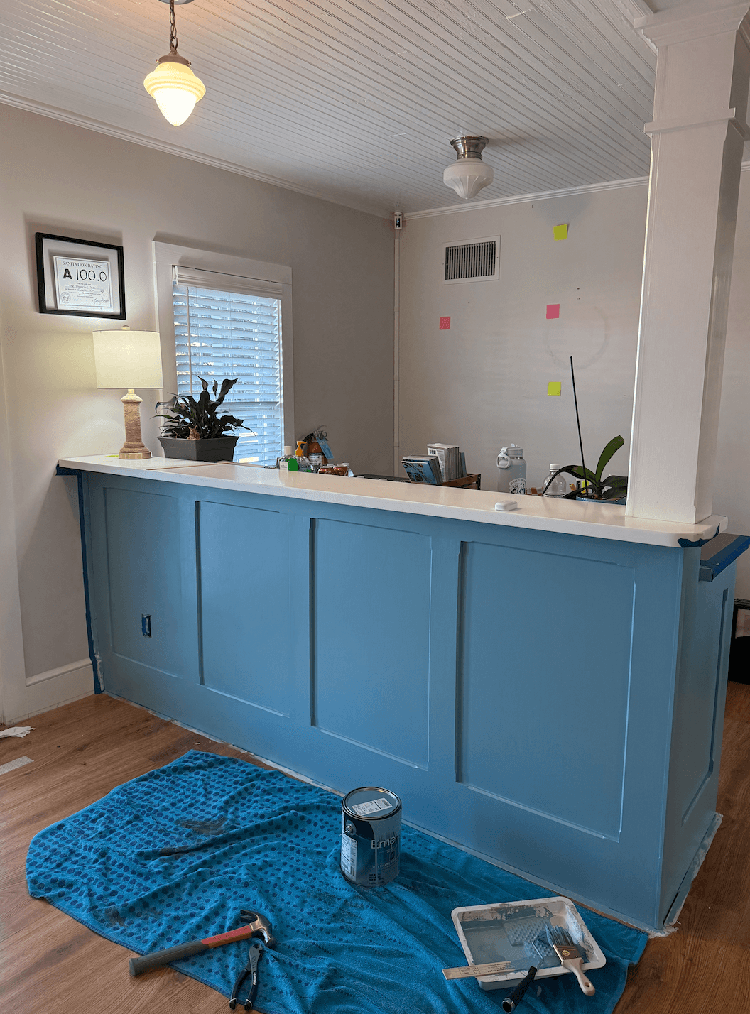 A partially painted blue reception desk with painting supplies and a lamp in a well-lit room.