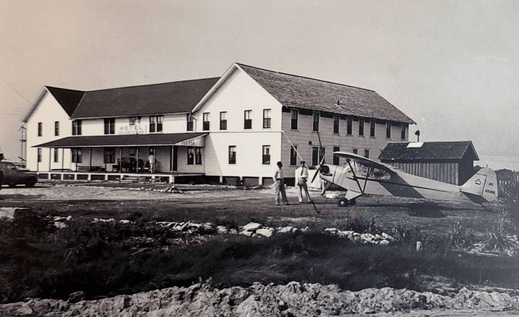 A black-and-white photo of a farmhouse with a parked airplane and people nearby.
