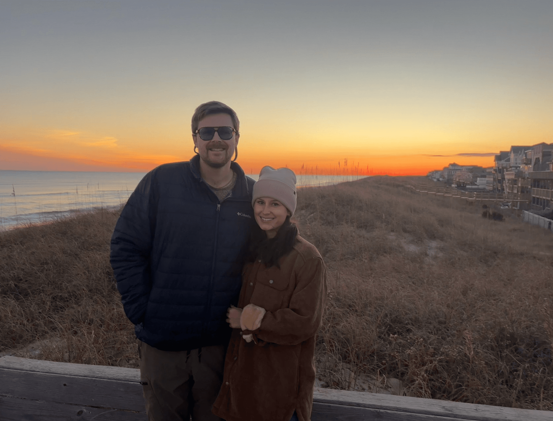 A couple stands together on a beach at sunset, with waves and beach houses in the background.