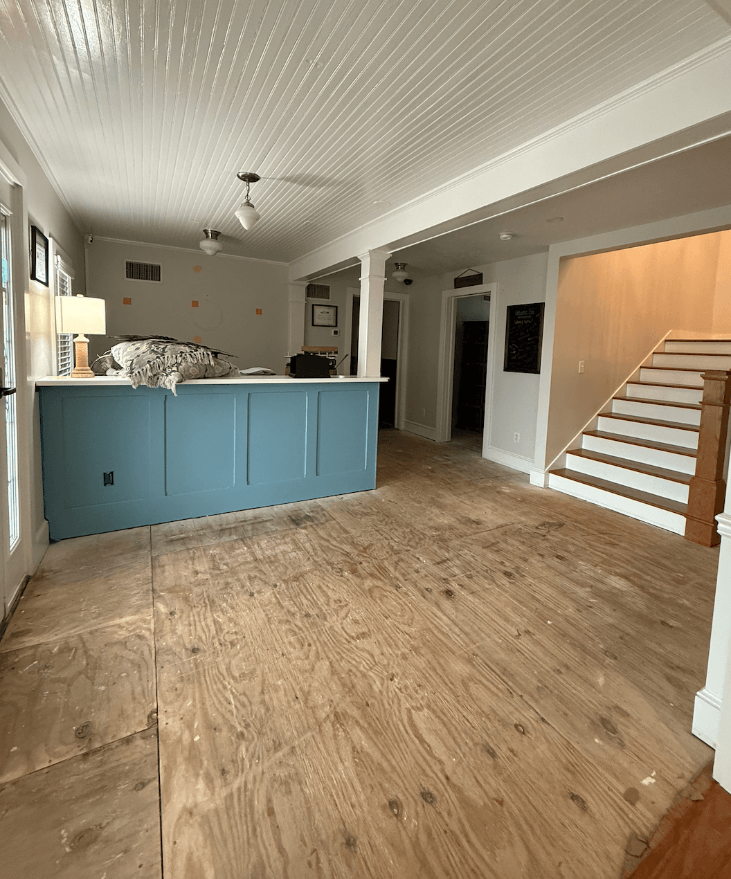 A spacious, unfinished living area featuring a blue kitchen island, plywood flooring, and a staircase leading to an upper level.