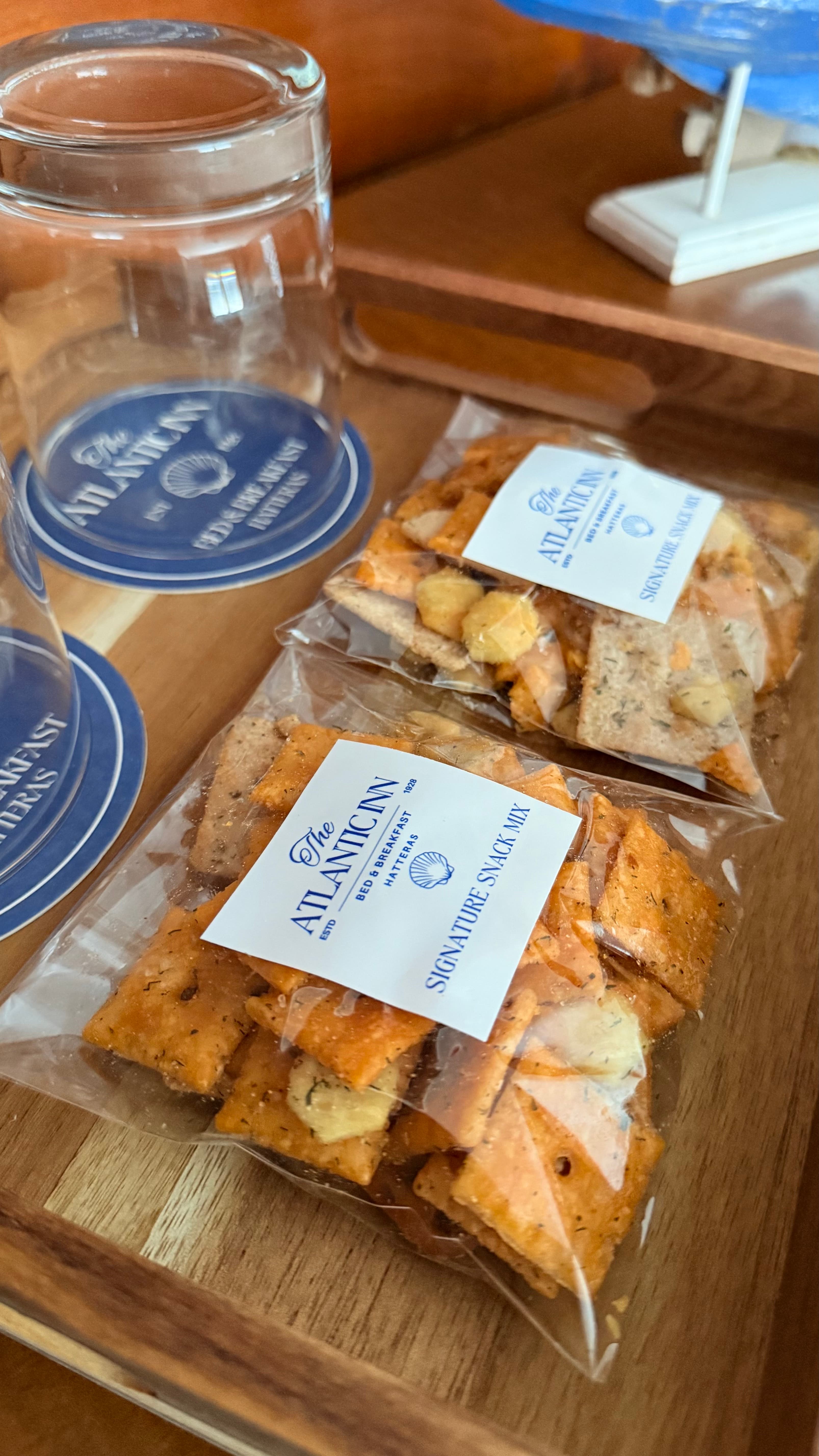 A packaged snack mix labeled "The Atlantic Inn Signature Snack Mix" beside coasters and glass jars on a wooden surface.