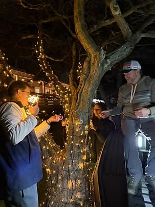 A group of three people enjoy a festive atmosphere around a tree adorned with lights at night.