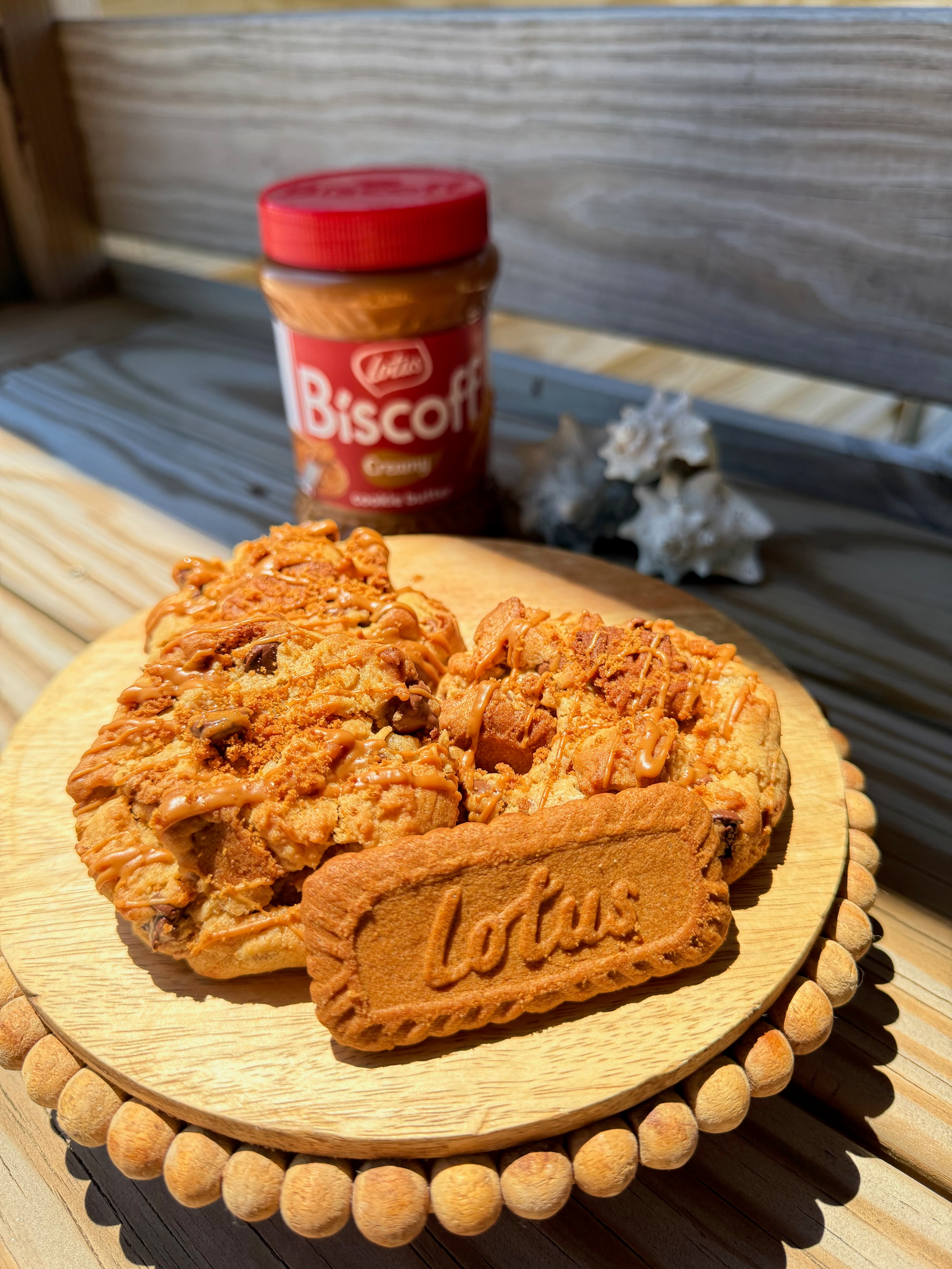 A wooden plate holds two cookies topped with Biscoff spread and a Lotus biscuit, with a jar of Biscoff spread in the background.