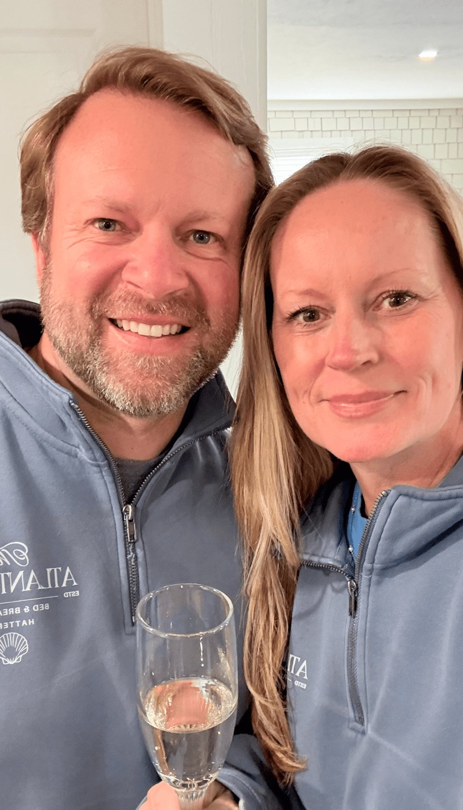 A smiling couple poses for a selfie, holding a champagne flute, both wearing matching gray jackets.