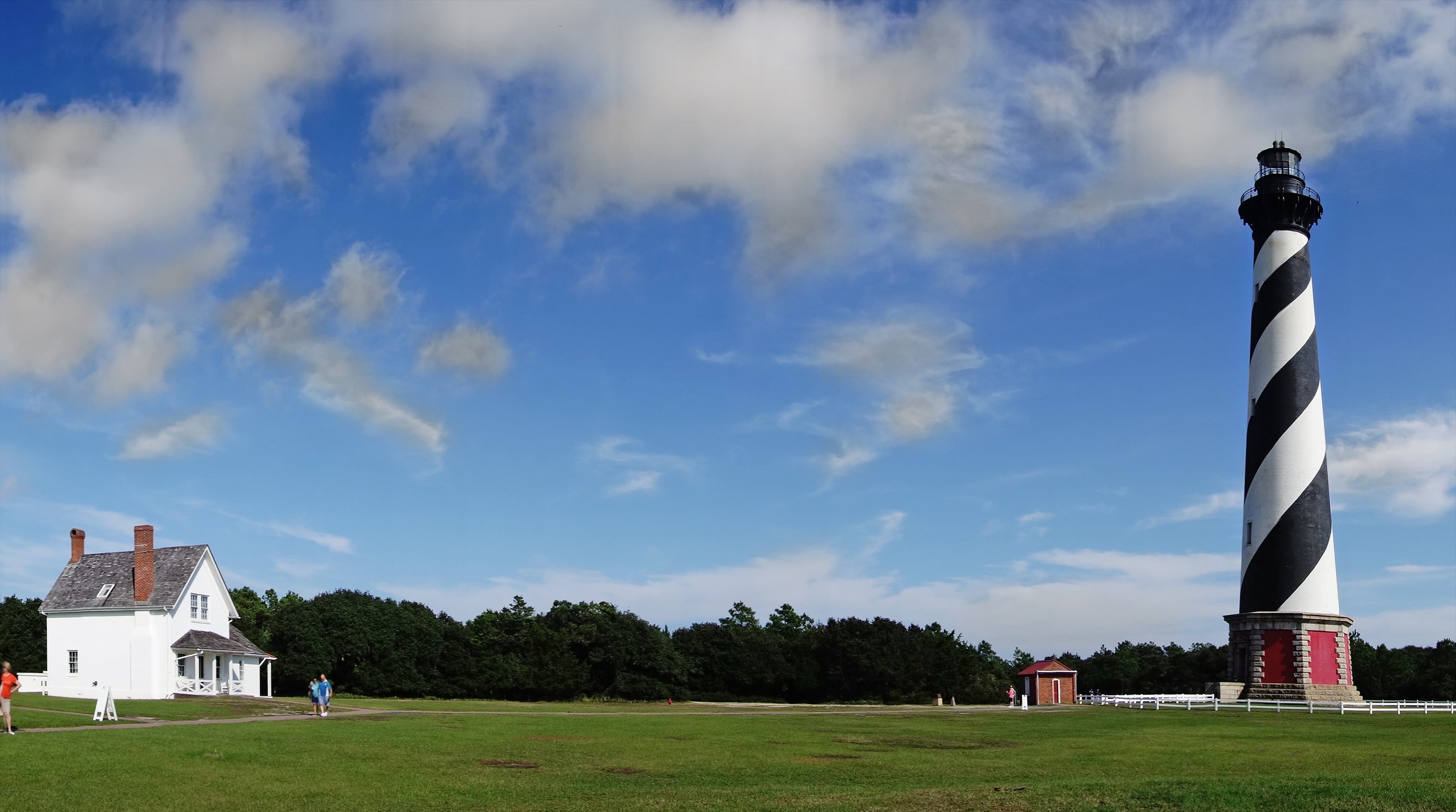 A black-and-white striped lighthouse stands beside a white house against a blue sky with scattered clouds.