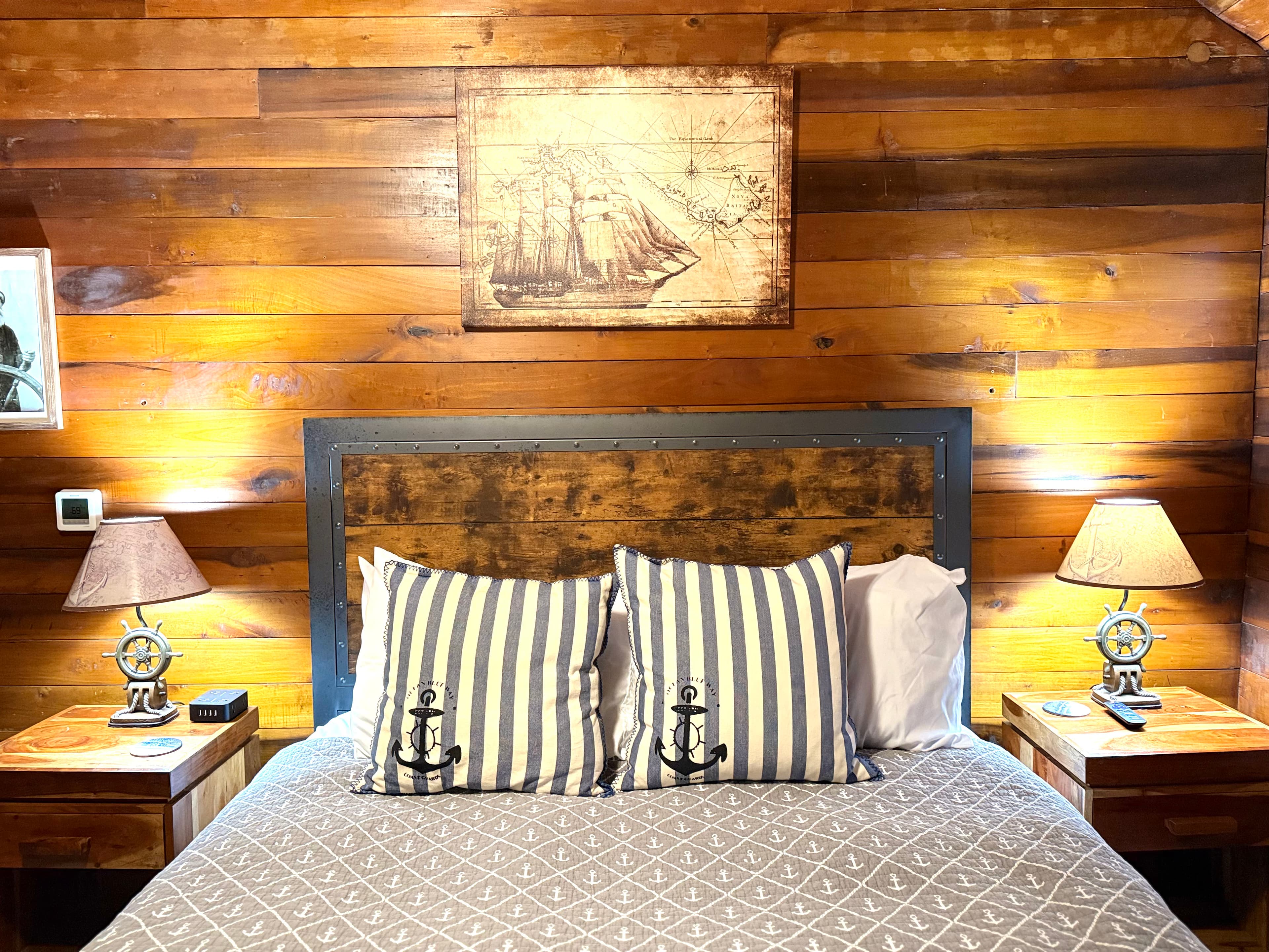 Cozy bedroom with wooden walls, striped anchor-themed pillows, and nautical decor.
