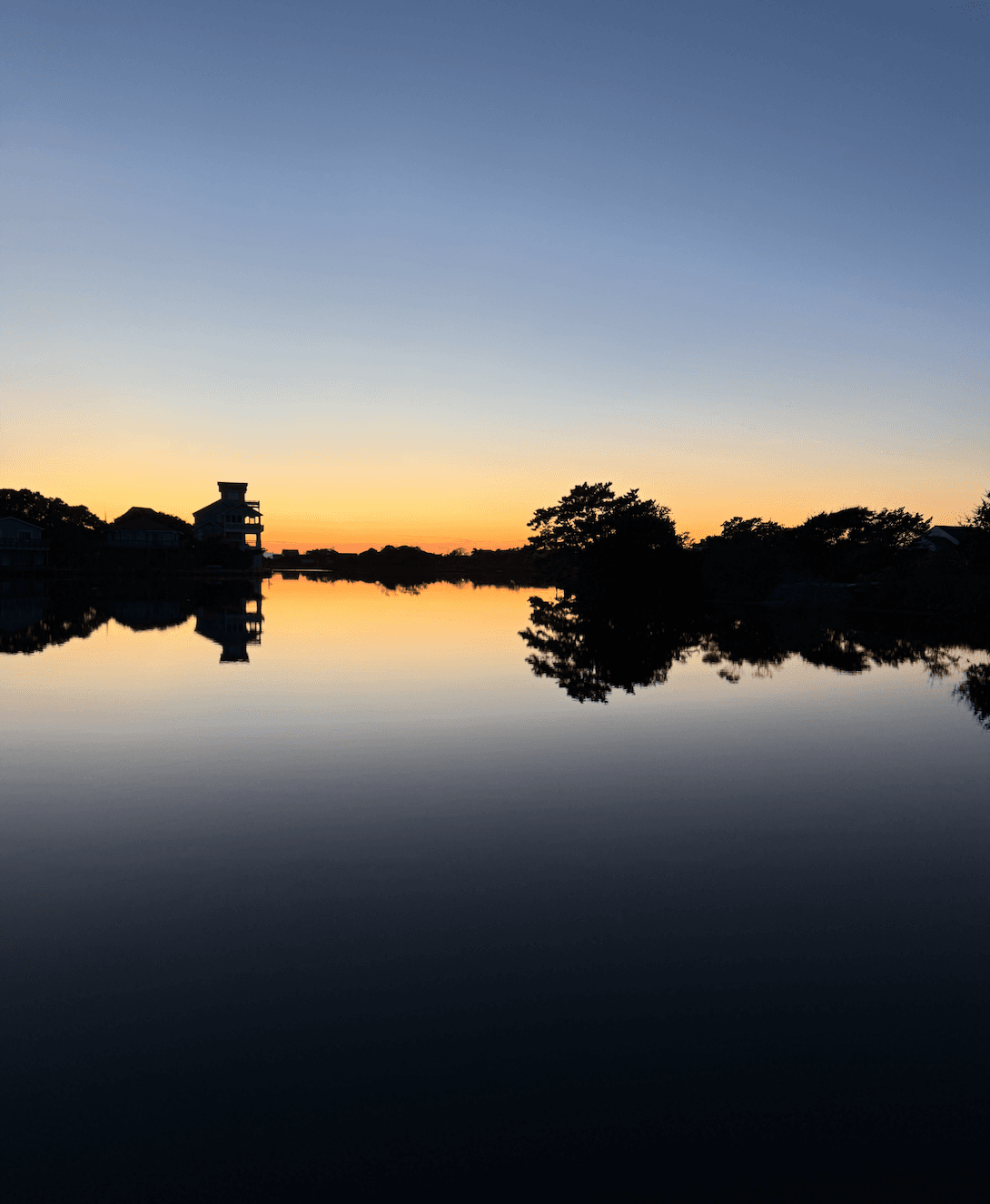 Silhouette of trees and a building reflected in a calm lake at sunset.