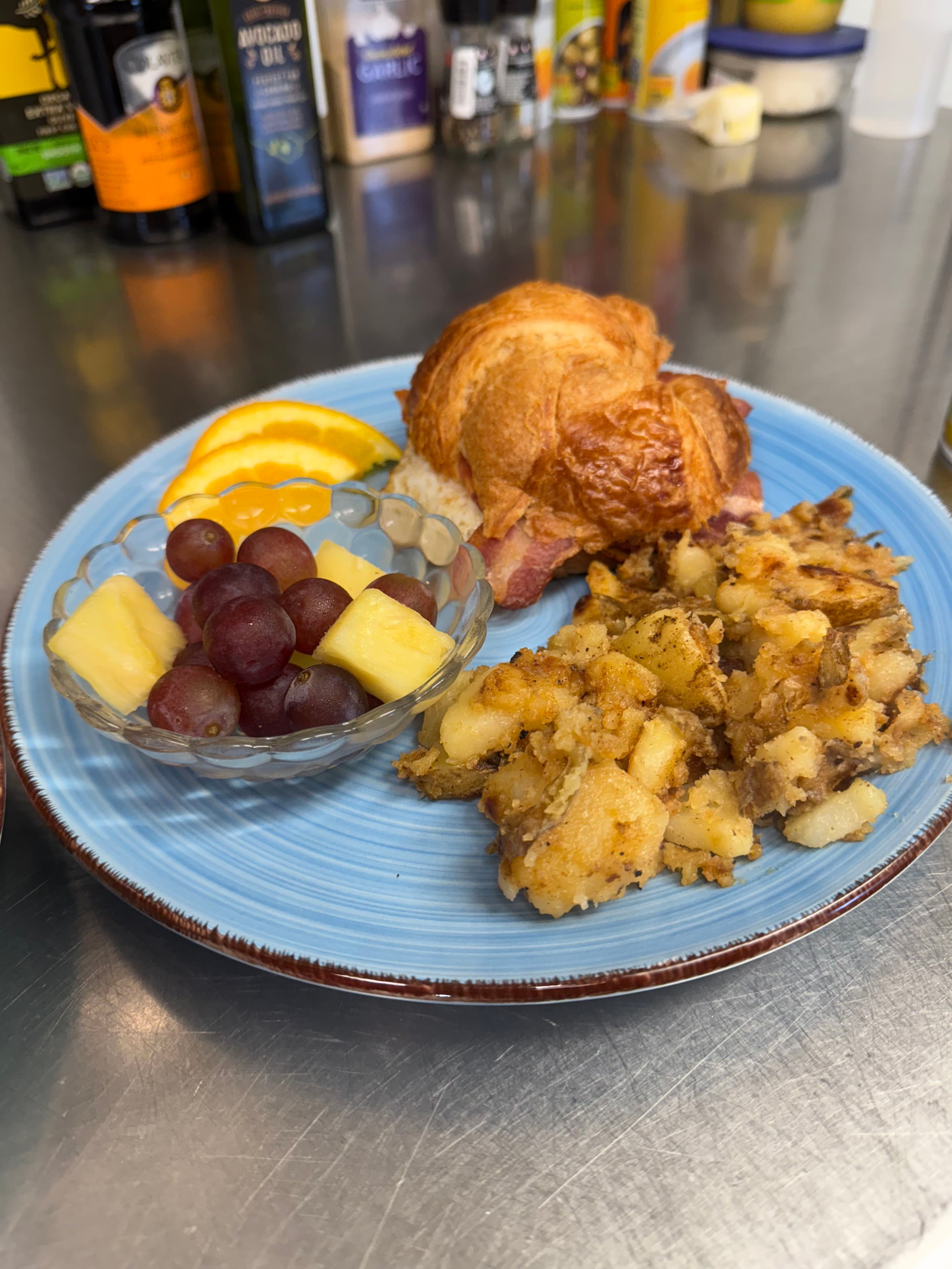 A plate featuring a croissant with bacon, sliced oranges, a bowl of grapes and pineapple, and seasoned potato hash.