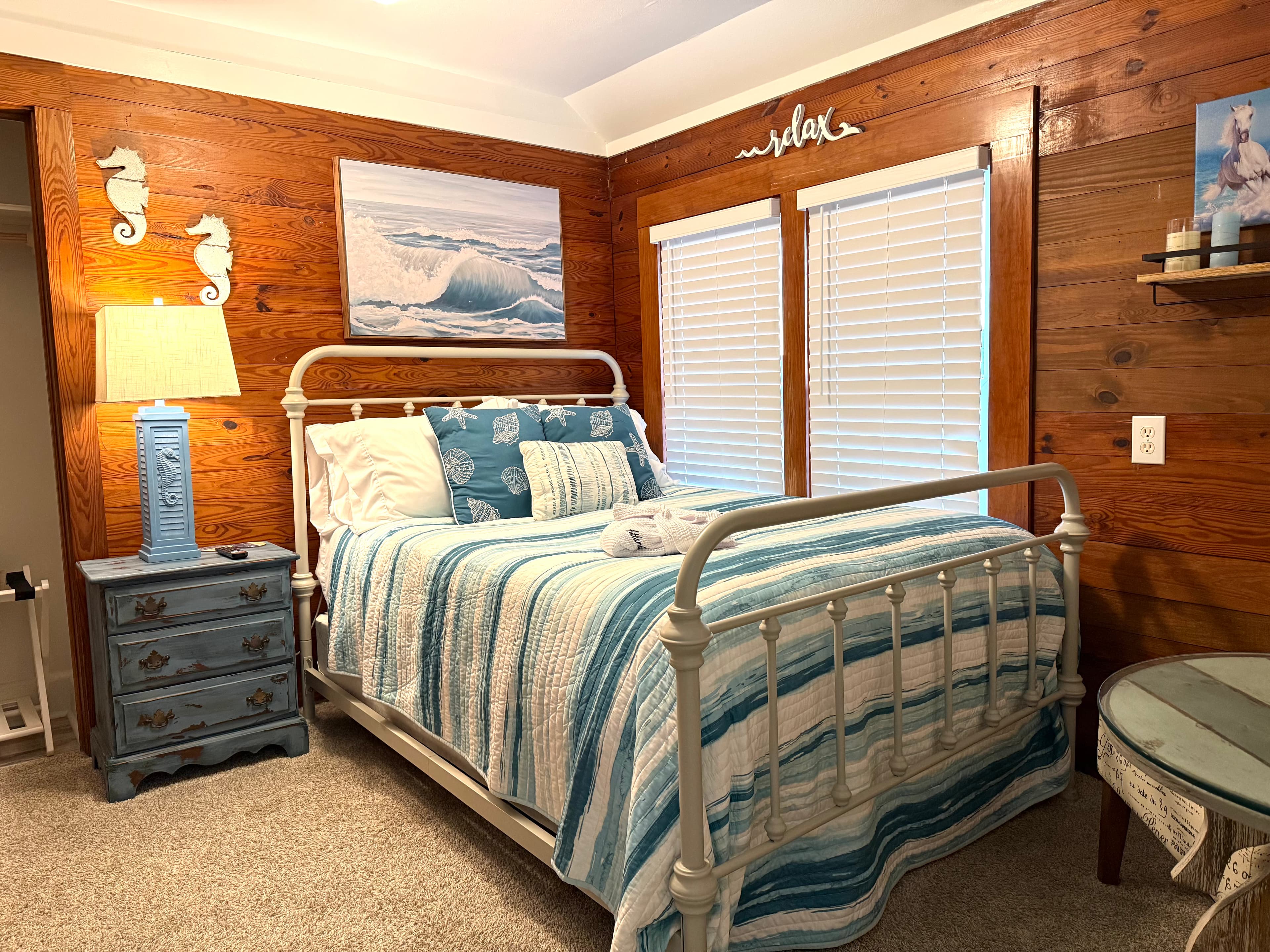 A cozy bedroom featuring a metal bed frame with blue and white bedding, a bedside table, and wooden walls adorned with beach-themed decor.