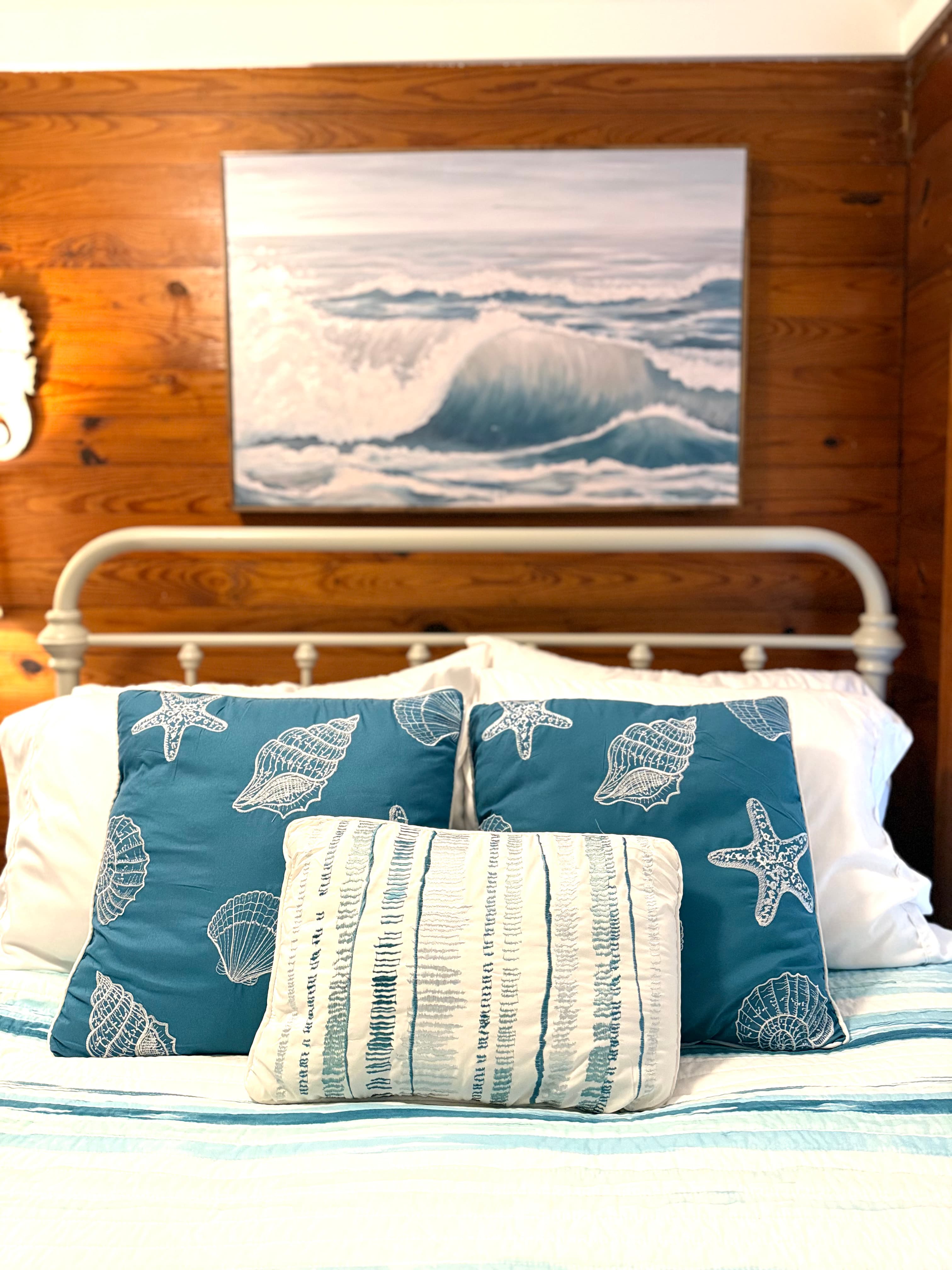 Cozy bed with blue shell-patterned pillows and a wave painting on the wooden wall.
