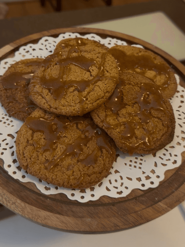 A wooden platter holds several caramel-drizzled cookies resting on a lace doily.