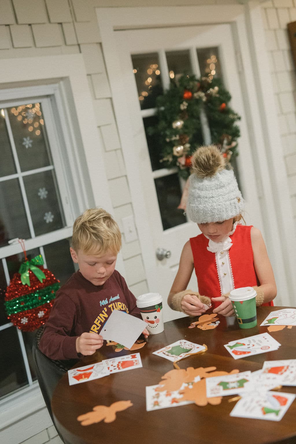 Two children are crafting holiday cards at a table decorated with cutouts and drinks, with festive decor in the background.