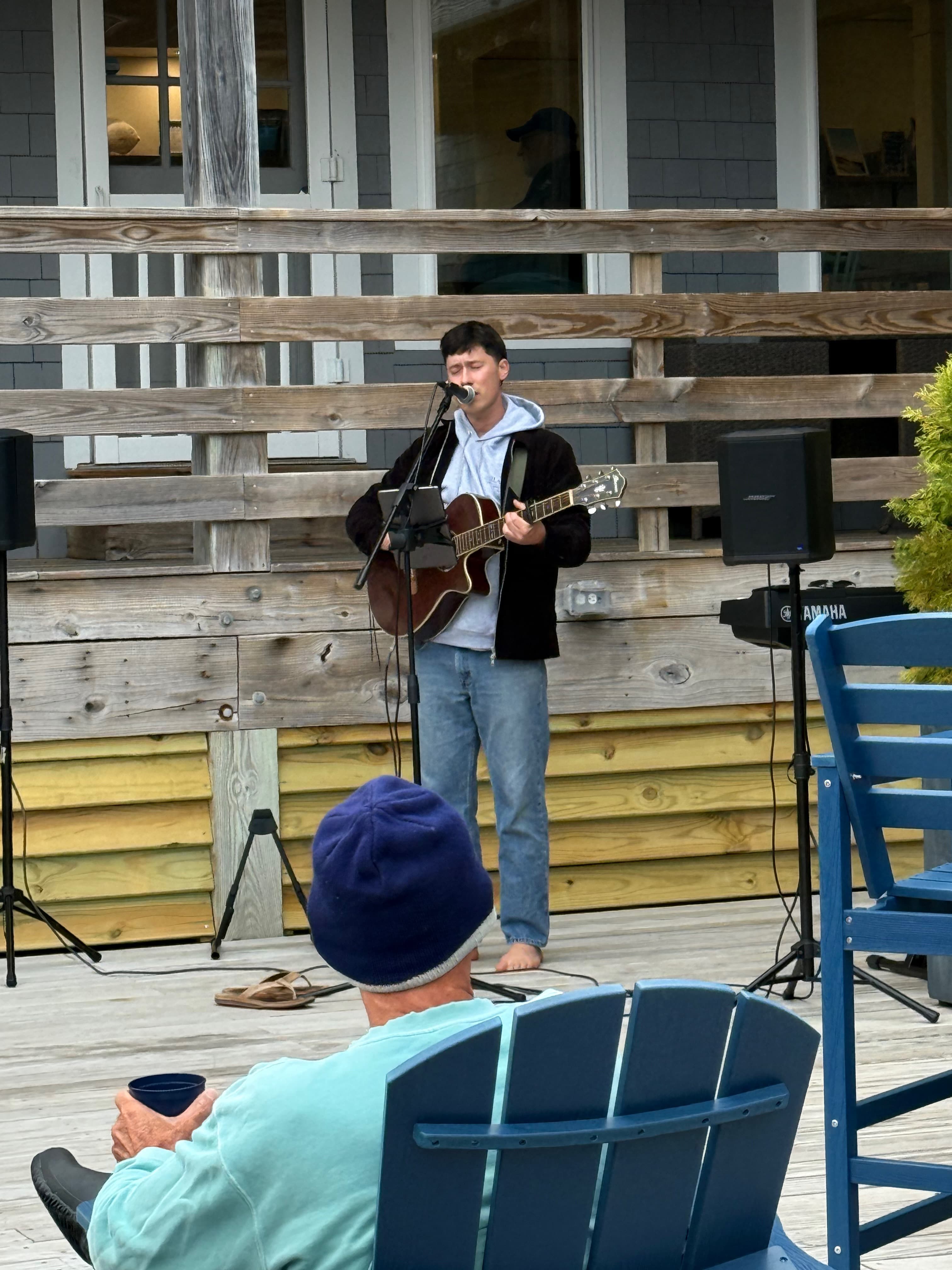 A young man plays guitar and sings on a wooden deck while an audience member sits in a chair, sipping a drink.